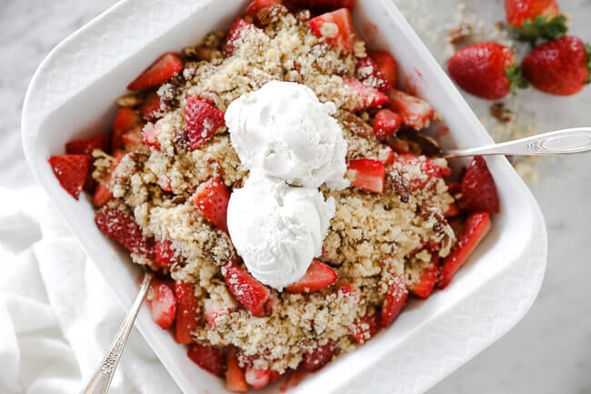 Horizontal overhead image of strawberry crumble in baking dish with two spoons dug in and two scoops of dairy-free ice cream on top. 