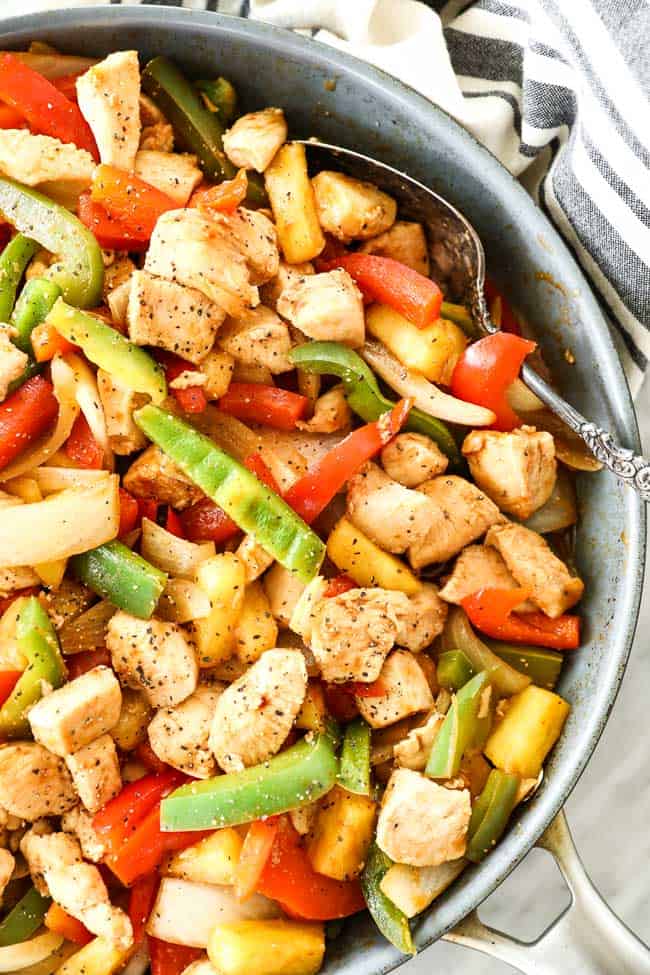 Overhead close up shot of healthy sweet and sour chicken in skillet with serving spoon. Dish includes chicken, onion, red and green bell pepper, pineapple and a sweet and sour sauce. 