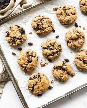 Overhead vertical image of a baking sheet with healthy vegan oatmeal raisin cookies. Extra oats and raisins in ramekins on the side.
