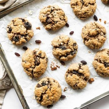 Overhead vertical image of a baking sheet with healthy vegan oatmeal raisin cookies. Extra oats and raisins in ramekins on the side.