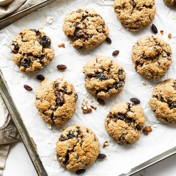 Overhead vertical image of a baking sheet with healthy vegan oatmeal raisin cookies. Extra oats and raisins in ramekins on the side.