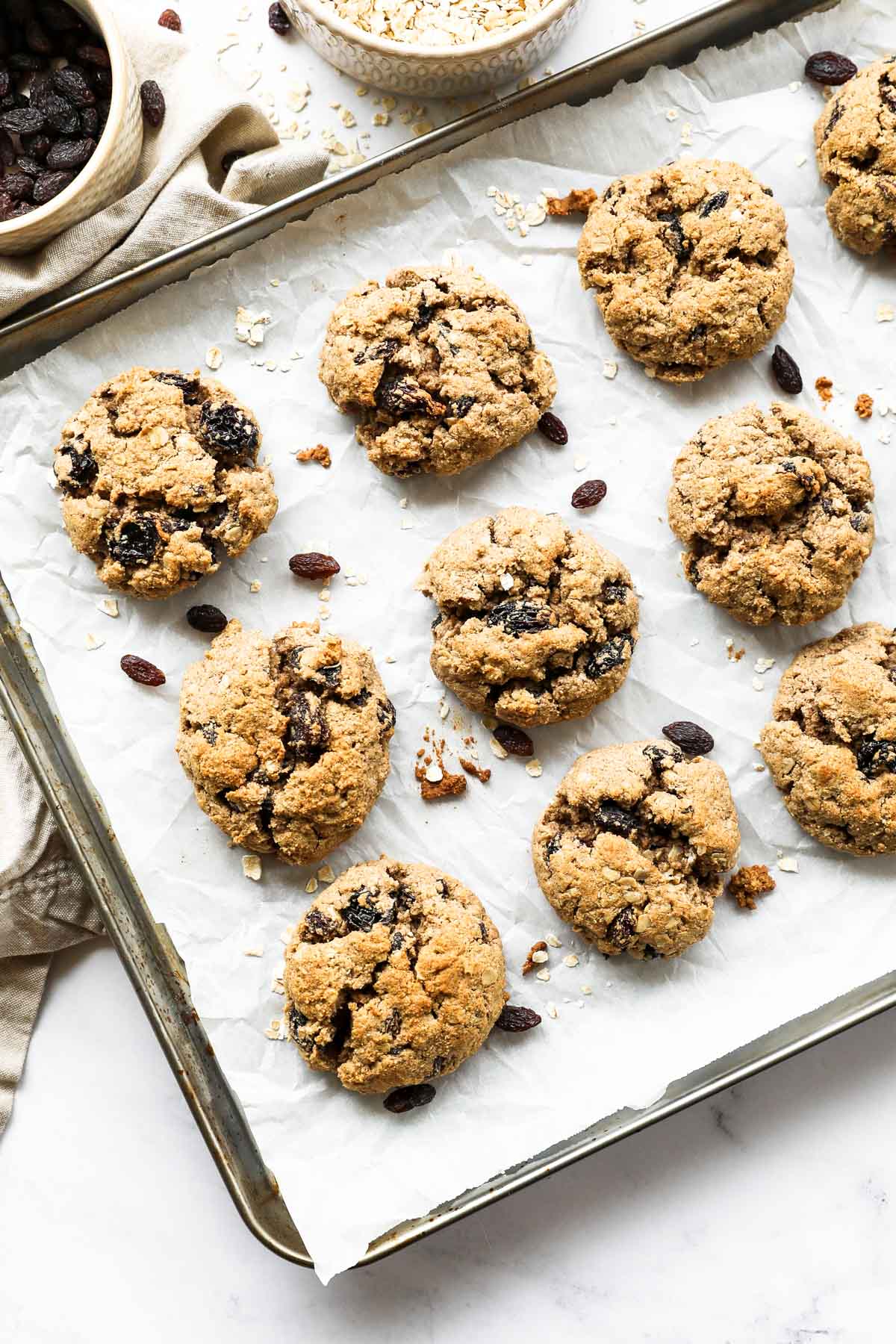 Overhead vertical image of a baking sheet with healthy vegan oatmeal raisin cookies. Extra oats and raisins in ramekins on the side. 