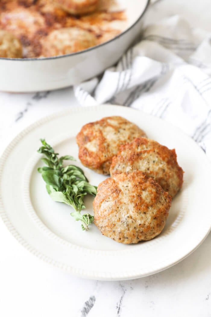 Angled image of three chicken sausage patties on a plate with fresh oregano on the side.