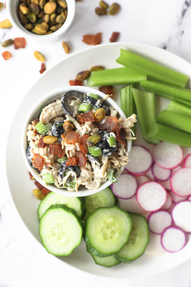 Vertical overhead image of chicken salad in a bowl on a plate with cucumber slices, radish slices and celery sticks. 