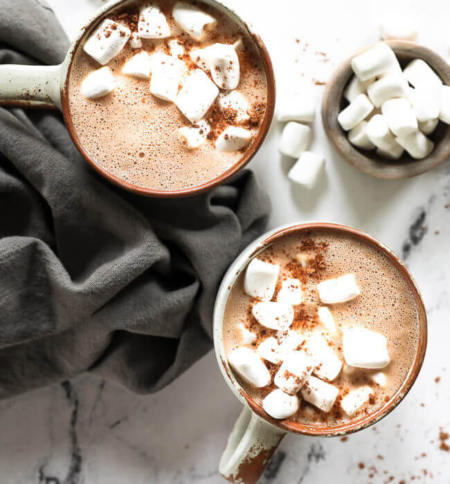 Overhead image of two mugs of vegan hot chocolate with marshmallows and cocoa powder on top.