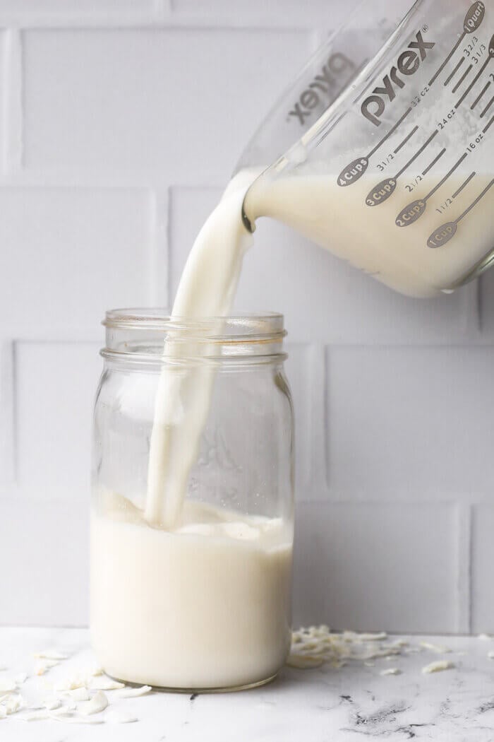 Pouring strained coconut milk into a mason jar for storing in the fridge.