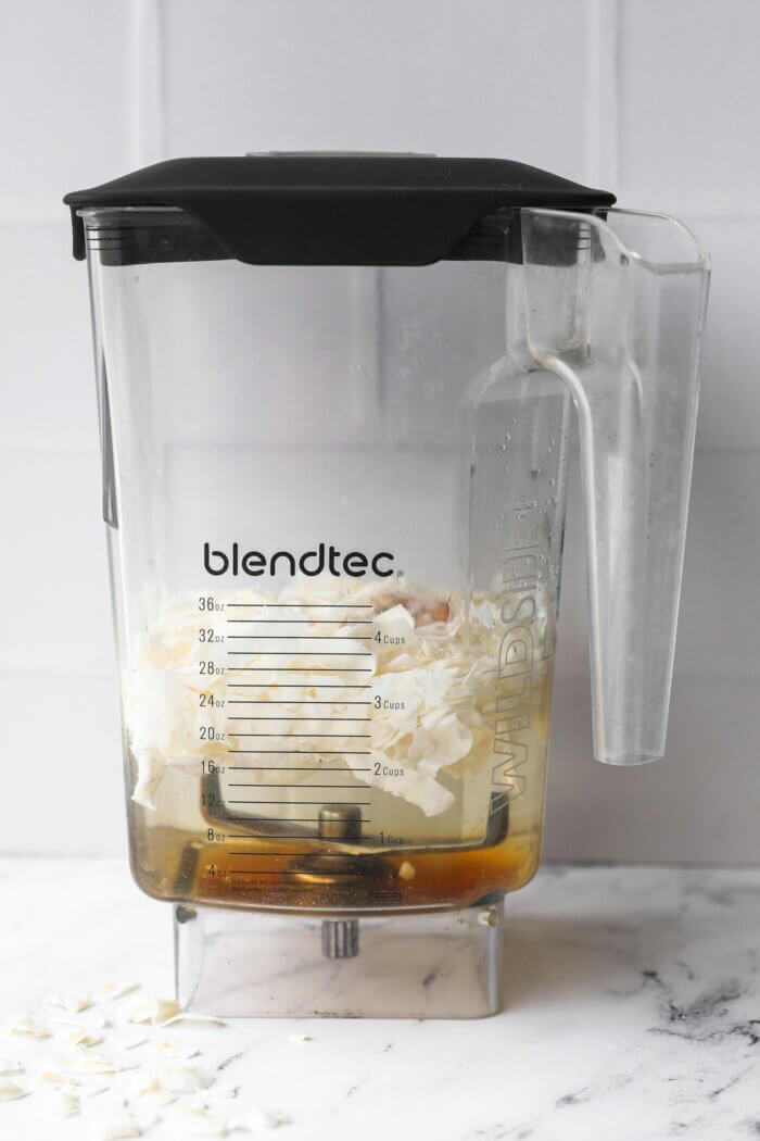 Straight on image of a blender with water, coconut flakes, maple syrup, vanilla and cinnamon before blending.