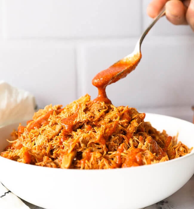 Bowl of shredded instant pot buffalo chicken with sauce pouring in from a spoon