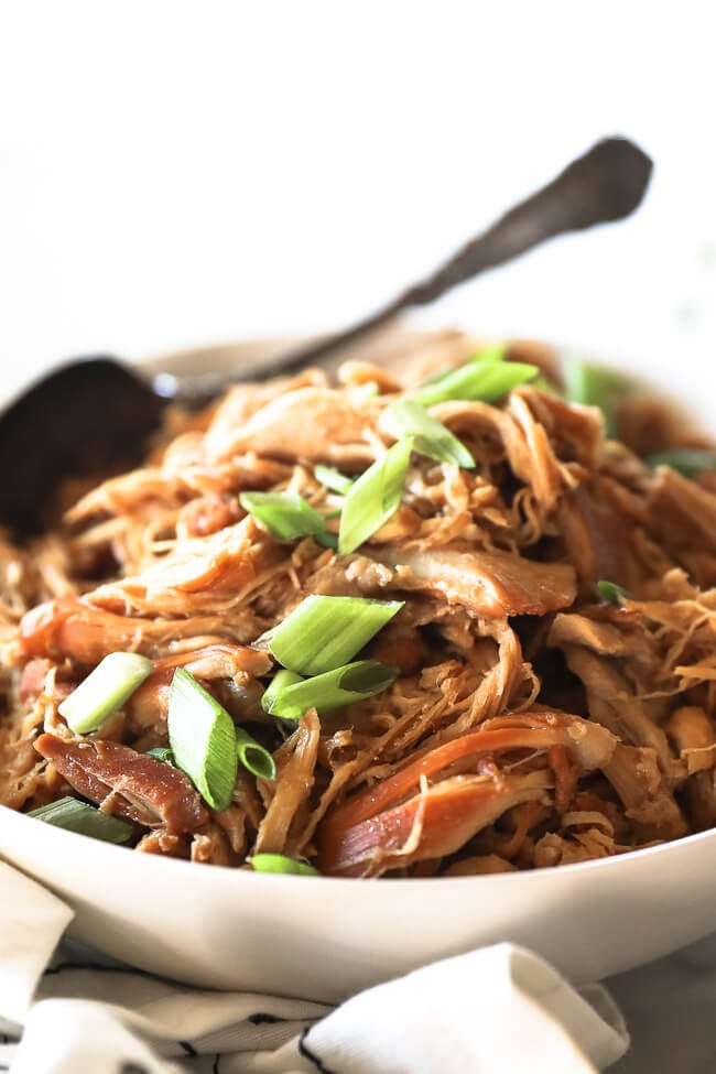 Angled close up image of instant pot honey garlic chicken in bowl with green onion on top and serving spoon dug into bowl in the background. 