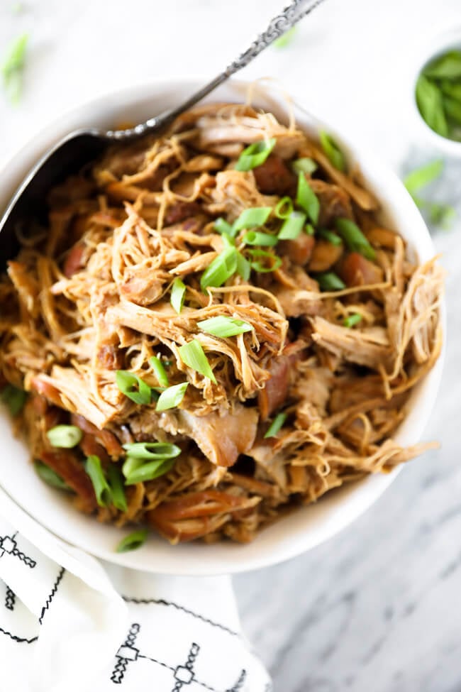 Close up overhead shot of honey garlic chicken in a bowl with chopped green onion sprinkled on top. Serving spoon is dug into bowl. 