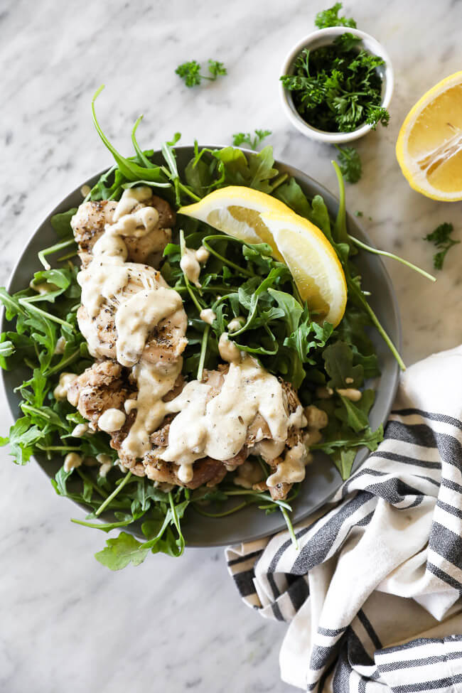 Chicken thighs covered in creamy sauce on top of arugula on a plate with lemon wedges vertical overhead image