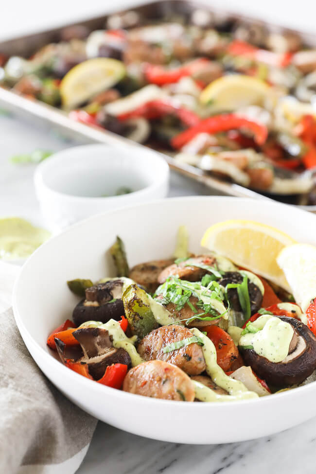 Vertical angled shot of italian sausage and peppers recipe served in a bowl with drizzle of herb aioli on top. Sheet pan in the background. 