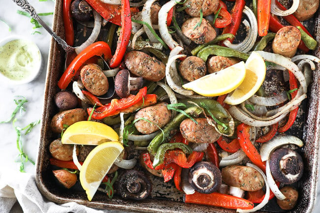 Horizontal overhead image of sheet pan filled with Italian sausage, bell peppers, onion and mushrooms. Garnished with fresh basil and lemon wedges. 