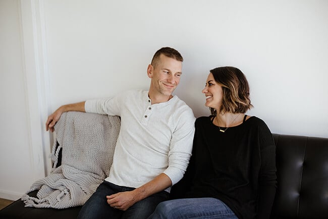 Horizontal image of Justin and Erica Winn sitting on couch smiling at each other. 