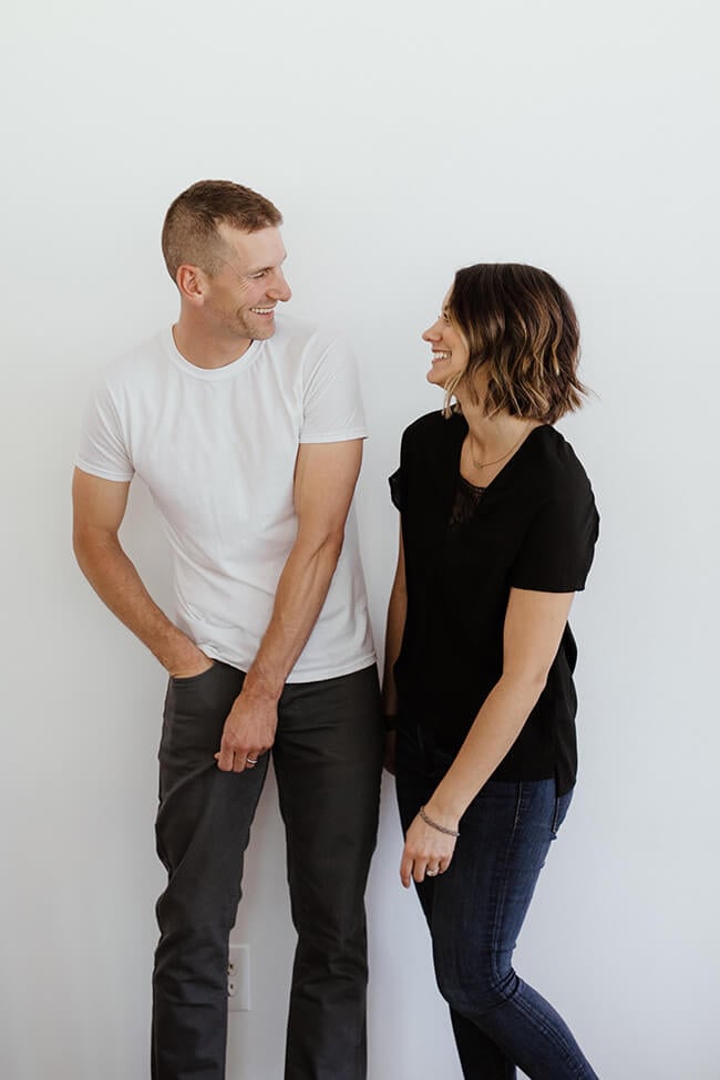 Vertical image of Justin and Erica Winn standing against a white wall leaning in toward each other laughing. 