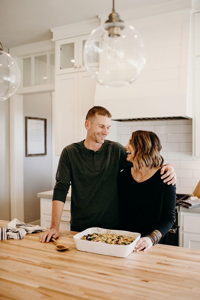 Justin + Erica Winn standing at kitchen counter with casserole dish smiling at each other