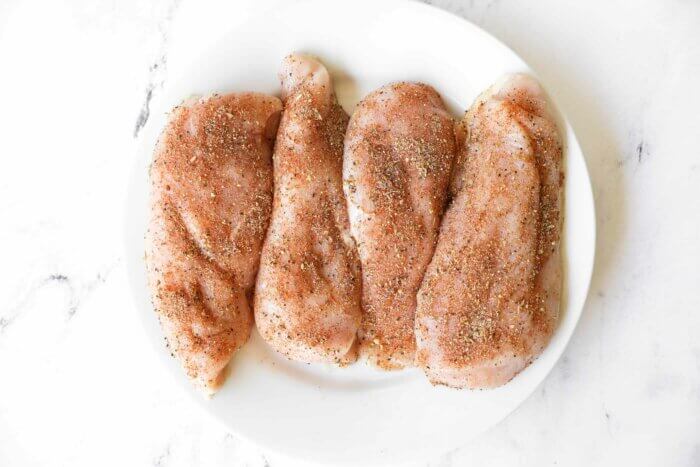 Four raw chicken breasts coated in seasoning mix on a plate.