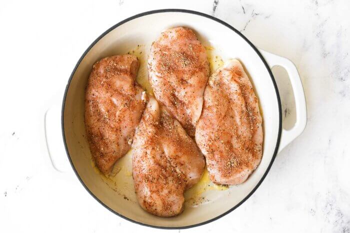 Four raw, seasoned chicken breasts ready to cook in a skillet with butter or ghee.
