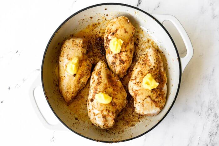 Browned chicken breasts in a skillet with a tablespoon of butter on top of each piece.