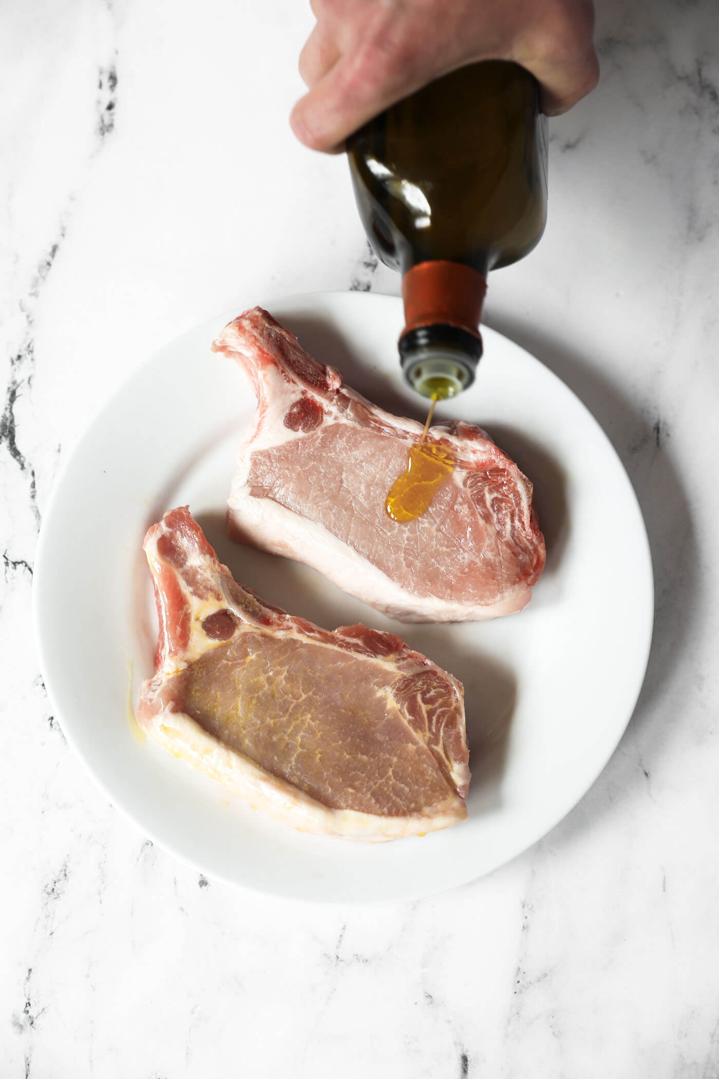 Pouring oil onto a pork chops before sprinkling seasoning on them.