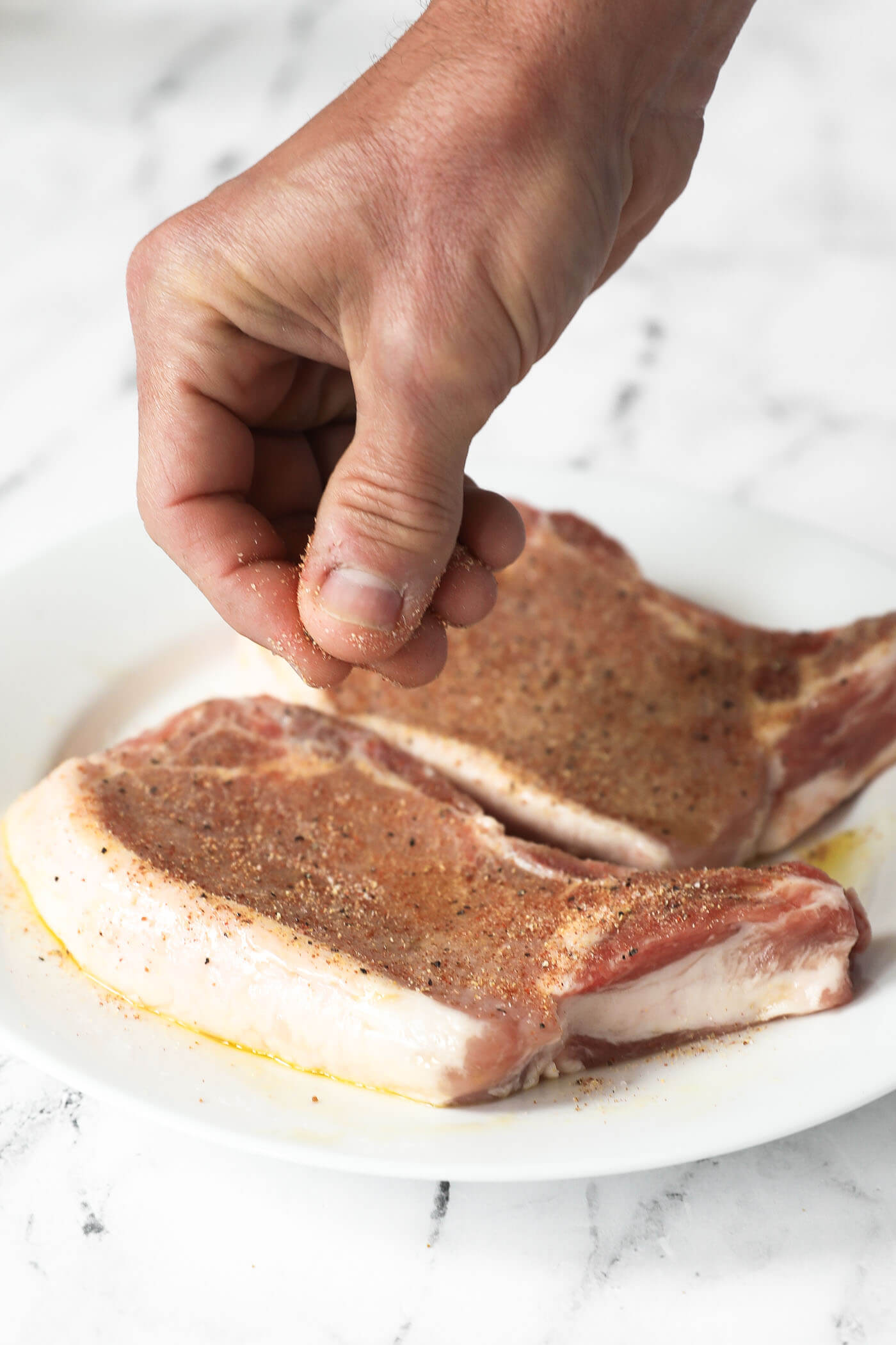 A hand sprinkling a seasoning mixture all over two pork chops.