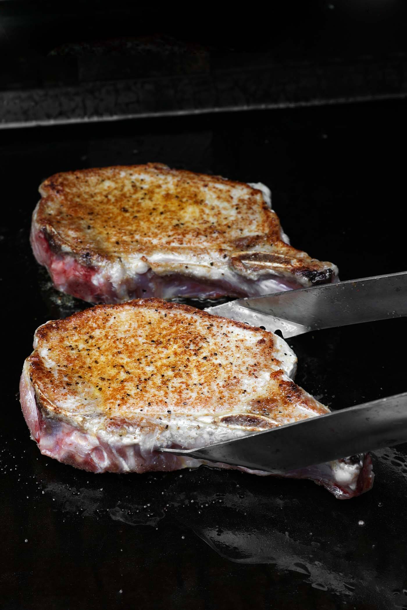 Two pork chops right after being flipped while they cook on a griddle.