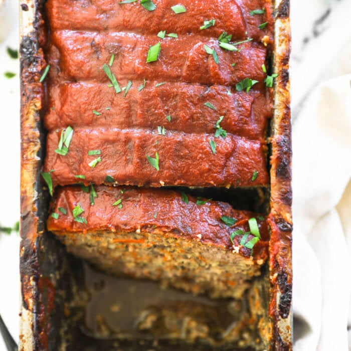 Overhead image of meatloaf in a loaf plan sliced and a couple pieces removed from the pan. The meatloaf is topped with paleo ketchup and chopped parsley.