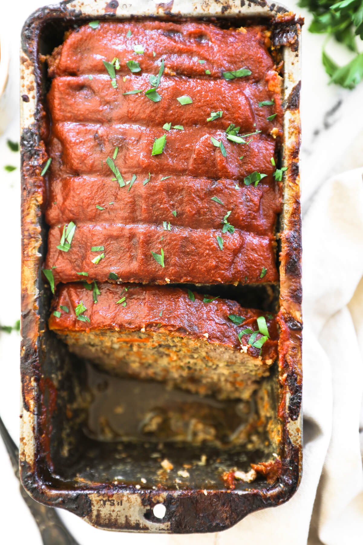 Overhead image of meatloaf in a loaf plan sliced and a couple pieces removed from the pan. The meatloaf is topped with paleo ketchup and chopped parsley.