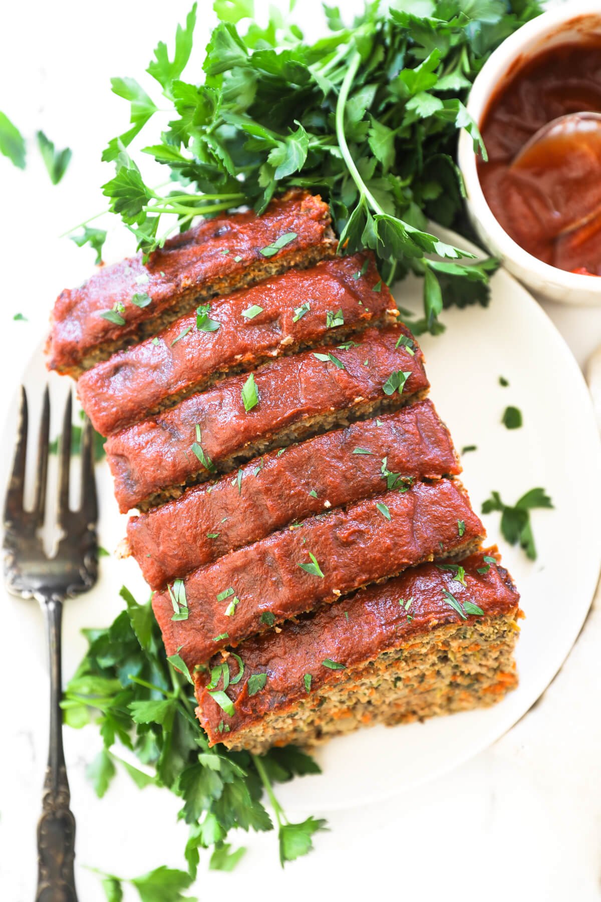 Overhead image of several pieces of meatloaf on a plate with a large fork and extra ketchup and parsley on the side.