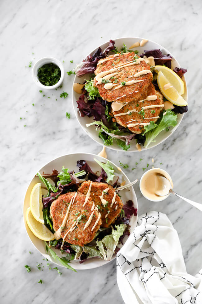 Vertical overhead image of two plates with two salmon patties on each plate served over fresh greens and drizzled with spicy aioli sauce. Extra sauce and chopped parsley on the side. 