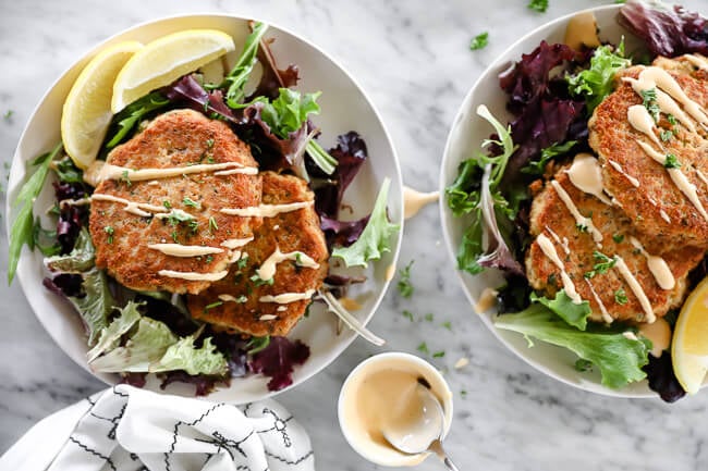 Horizontal overhead image of salmon patties on a plate served over fresh greens and topped with drizzle of spicy aioli sauce. 