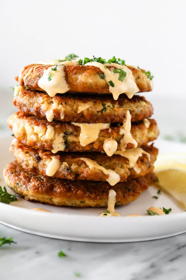 Close up vertical straight on shot of stack of salmon patties with sauce cascading down the sides. Chopped parsley sprinkled on top. 