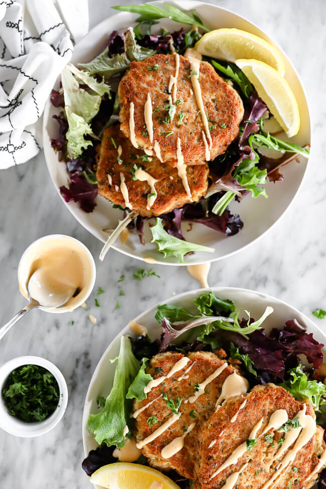 Vertical overhead image of two plates with two salmon patties on each plate served over fresh greens and drizzled with spicy aioli sauce. Extra sauce and chopped parsley on the side. 