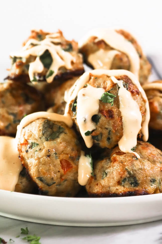 Vertical close up straight on angle image of keto meatballs stacked on a plate with spicy aioli sauce drizzled on top and cascading down. 