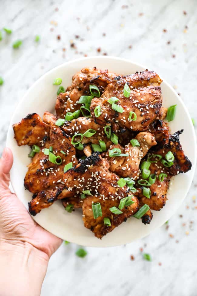Hand holding up Korean BBQ chicken on a plate with green onion slices.