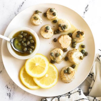 Lemon butter air fryer scallops on a white plate with butter sauce and capers on top. Lemon slices and capers in the side.