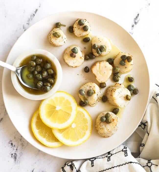 Lemon butter air fryer scallops on a white plate with butter sauce and capers on top. Lemon slices and capers in the side.