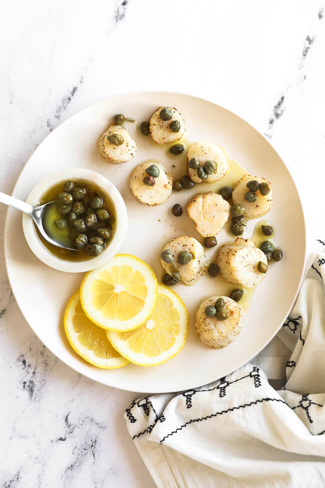 Air fryer scallops on a white plate with butter sauce and capers on top. Lemon slices and capers in the side.