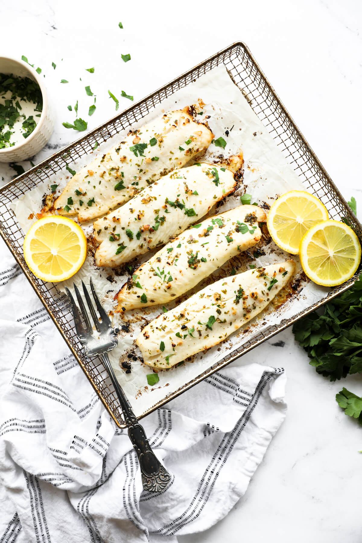 Overhead image of four pieces of tilapia in air fryer basket with sliced of lemon and fresh parsley to garnish.