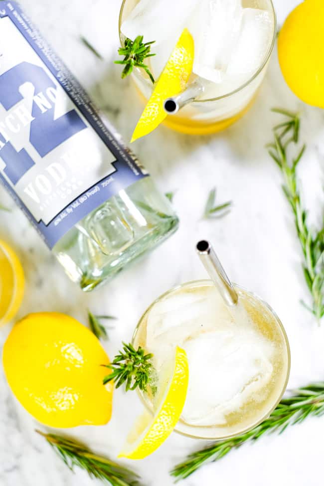 Two glasses of iced lemon & rosemary cocktail with lemon wedge, fresh rosemary garnish and stainless steel straws. Bottle of Heritage Distilling vodka. 