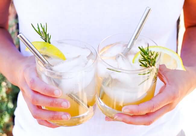 Holding two glasses of this lemon & rosemary cocktail with stainless steel straws in glasses. 