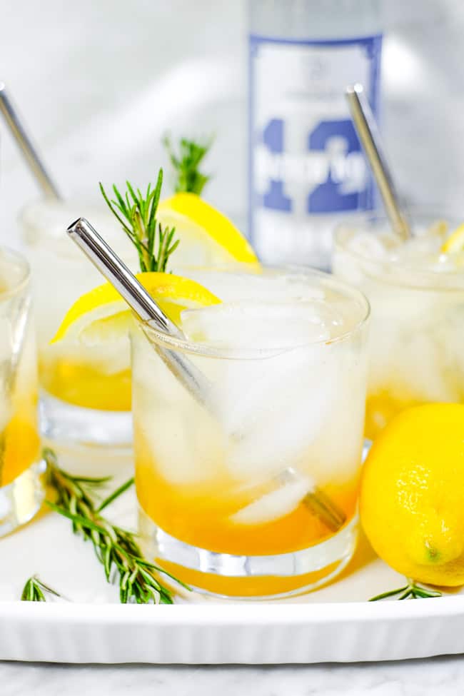 Tray with four glasses of iced lemon & rosemary cocktail with lemon wedges, fresh rosemary and stainless steel straws. 