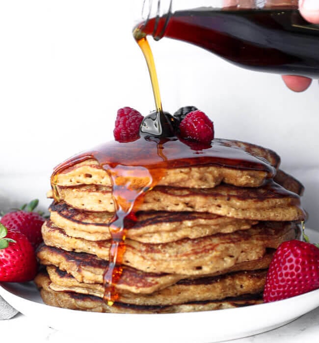 Stack of gluten free pancakes with maple syrup being poured on top and dripping down the side of the stack.
