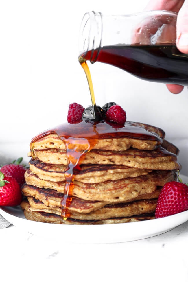 Straight on image of pouring maple syrup over a stack of gluten free pancakes with berries on top. 