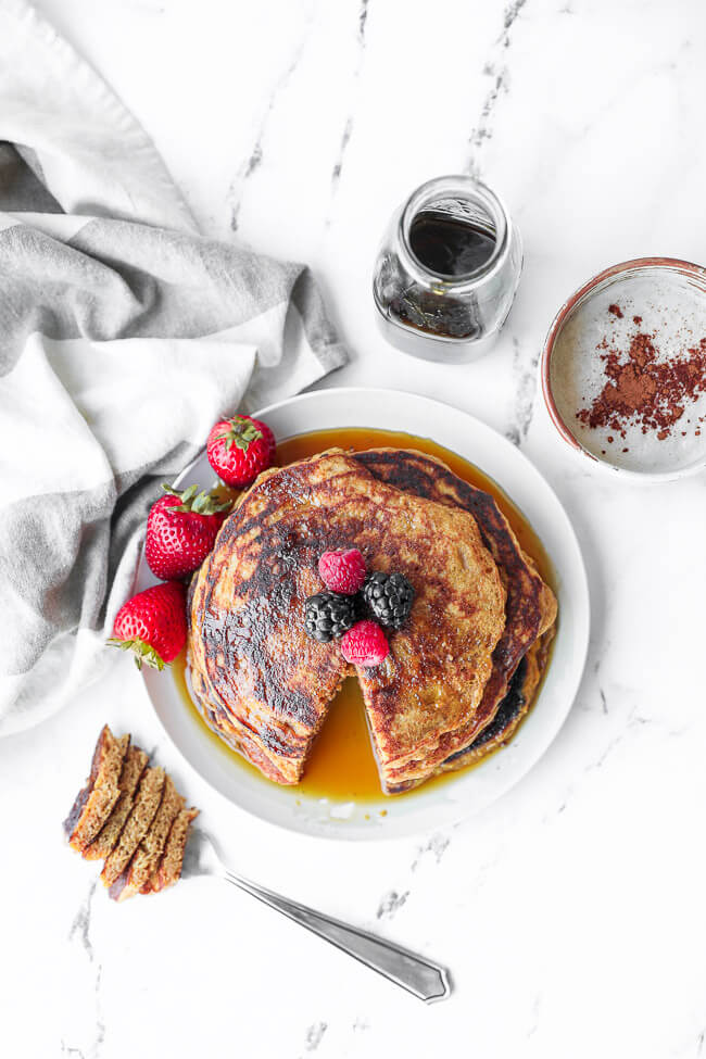 Overhead image of stack of vegan pancakes with a bite cut out. Berries on top with maple syrup drizzled on top. Extra syrup and a mug of coffee on the side. 