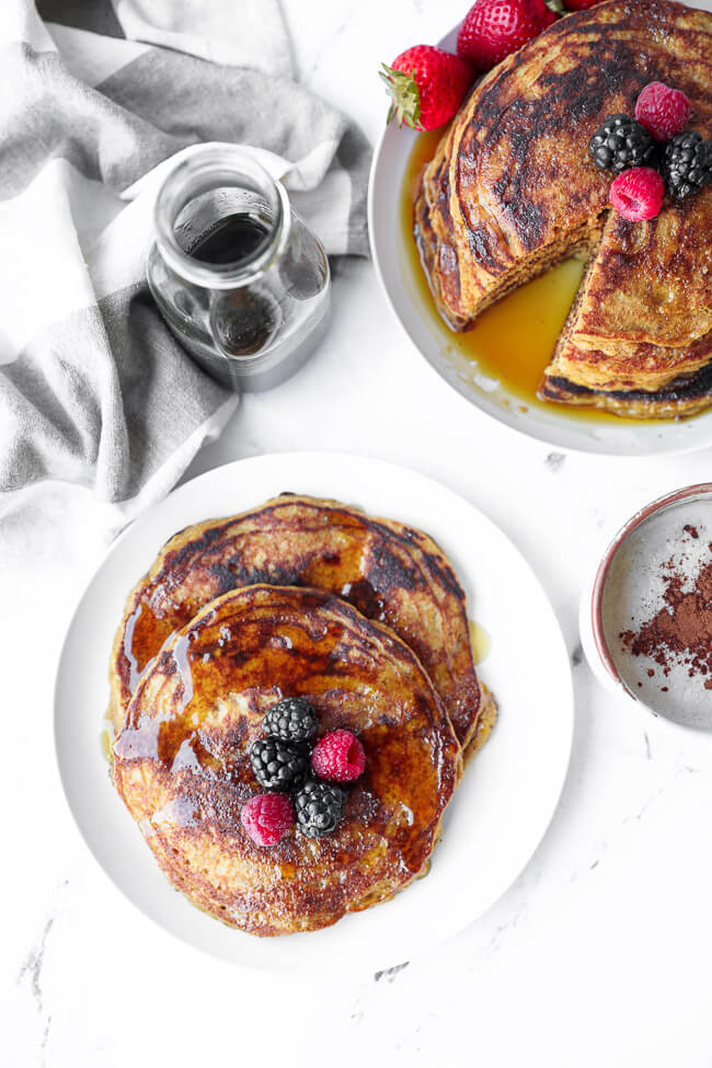 Overhead image of two gluten free pancakes on a plate with berries and maple syrup on top. Stack of pancakes, maple syrup and a mug of coffee off to the side. 