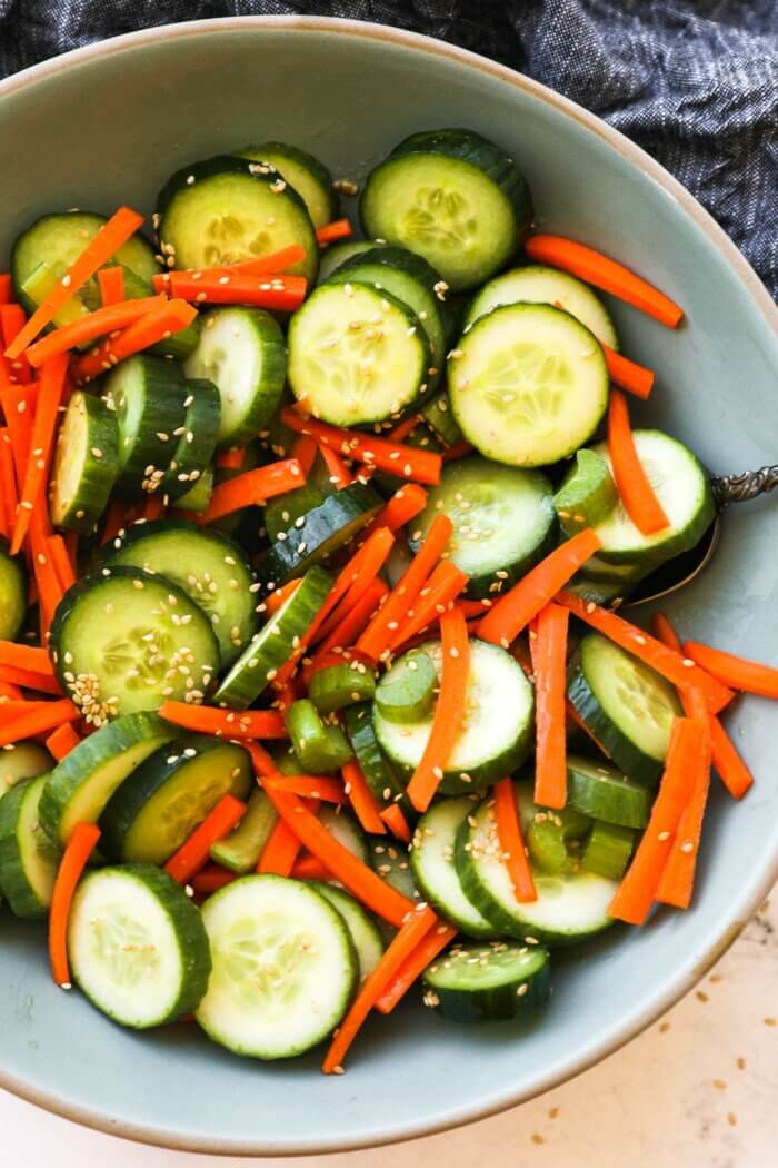 Close up overhead image of a bowl full of cucumber, carrots and celery with a sesame sauce and sesame seeds. Ingredients are all mixed up.