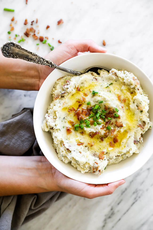 Vertical overhead image of holding a bowl of loaded mashed cauliflower with a serving spoon in it. 