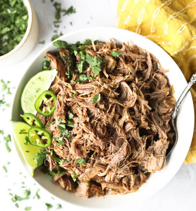Shredded marinated instant pot mojo pulled pork in a white bowl with lime, cilantro and jalapeno garnish