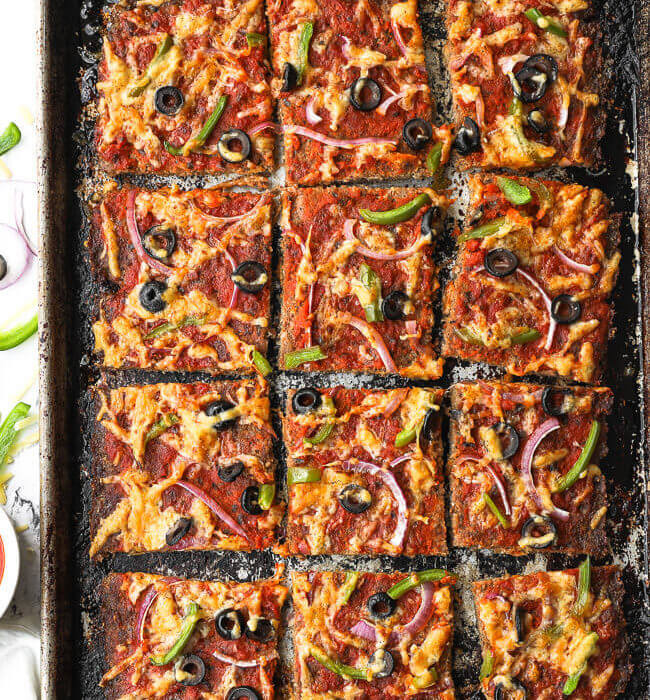 Overhead shot of sheet pan of meat pizza sliced into squares with cheese, olive, bell pepper and red onion topping